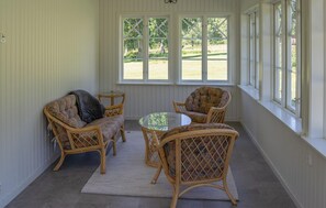 Un porche cubierto con mobiliario de mimbre, una mesa de vidrio y amplias ventanas con vista a un jardín.