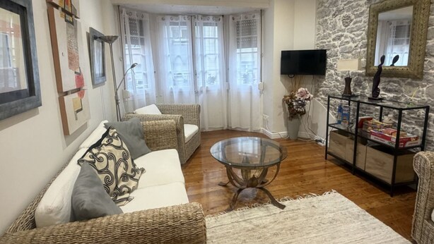 Uma sala de estar com sofá, mesa de centro de vidro, cadeiras de vime, parede de pedra, televisão e um espelho.