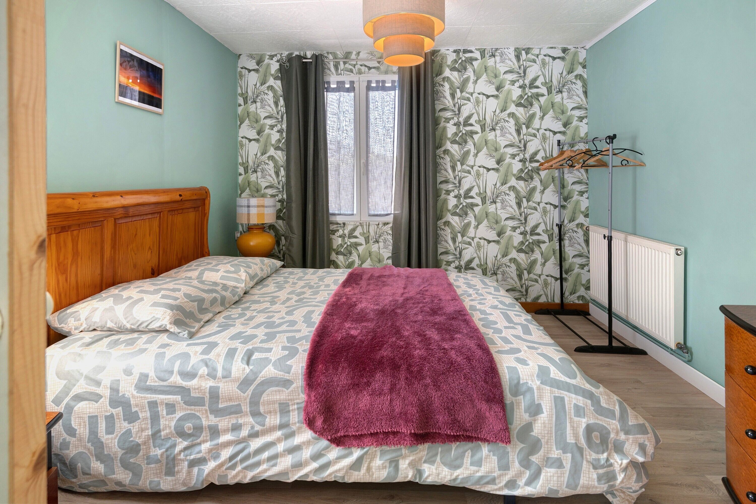 Une chambre à coucher avec un lit, une tête de lit en bois, un radiateur et une fenêtre avec des rideaux.