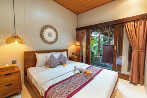 A hotel room with a bed, wooden headboard, two bedside lamps, a wall-mounted decorative piece, and a window with curtains. - Ubud Heritage Stay (Gianyar)