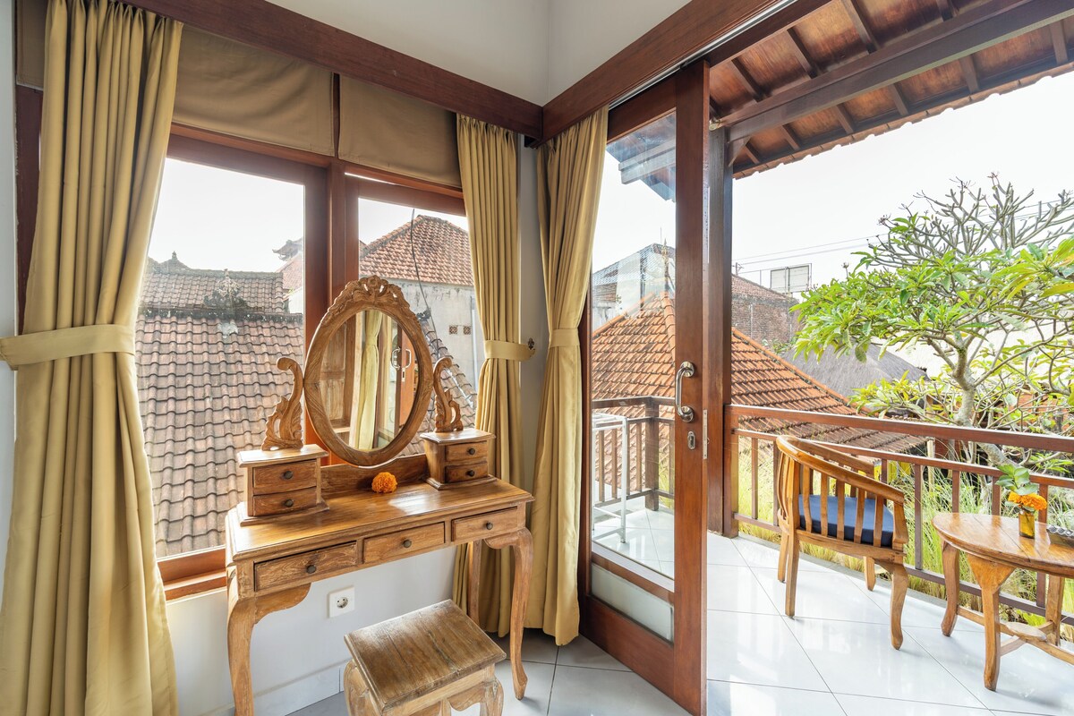 A room with a wooden vanity, a large mirror, and a small stool. There is a balcony with a wooden railing and a view of the neighborhood.