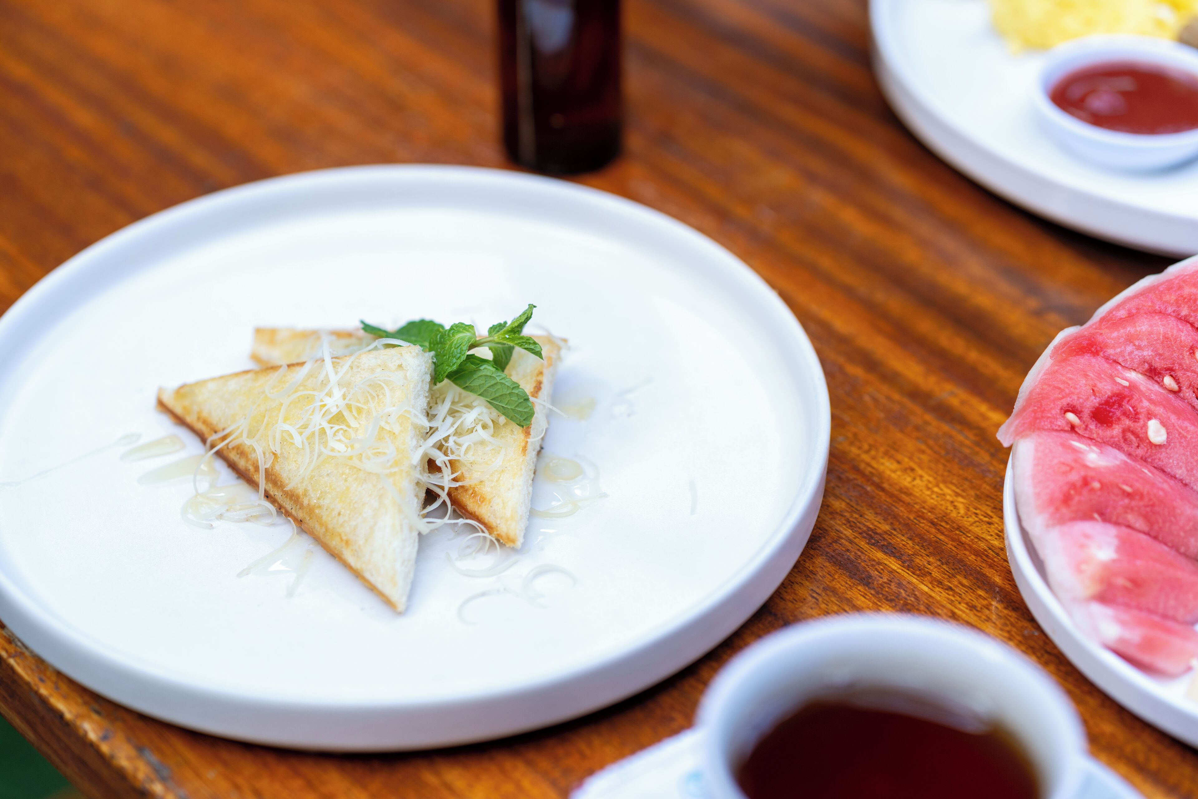 A plate with a triangular food item, possibly a pastry, garnished with cheese and mint leaves, accompanied by a small bowl of sauce and a slice of watermelon.
