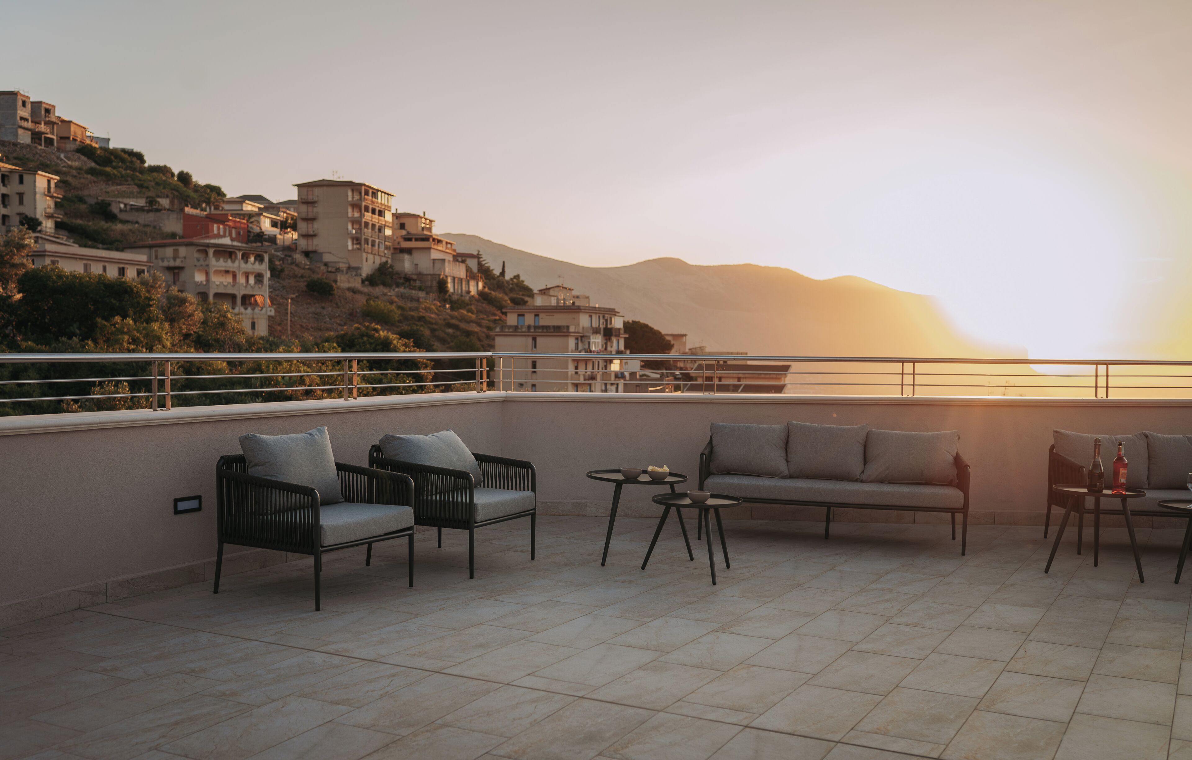 A terrace with outdoor furniture, a view of buildings on a hillside, and a sunset in the background.