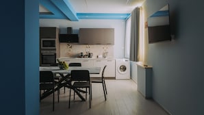 A compact kitchen with a dining area, a washing machine, and a flat-screen TV mounted on the wall.