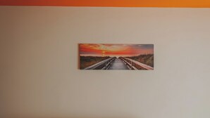 A beige sofa against a wall with a painting of a sunset over a bridge.