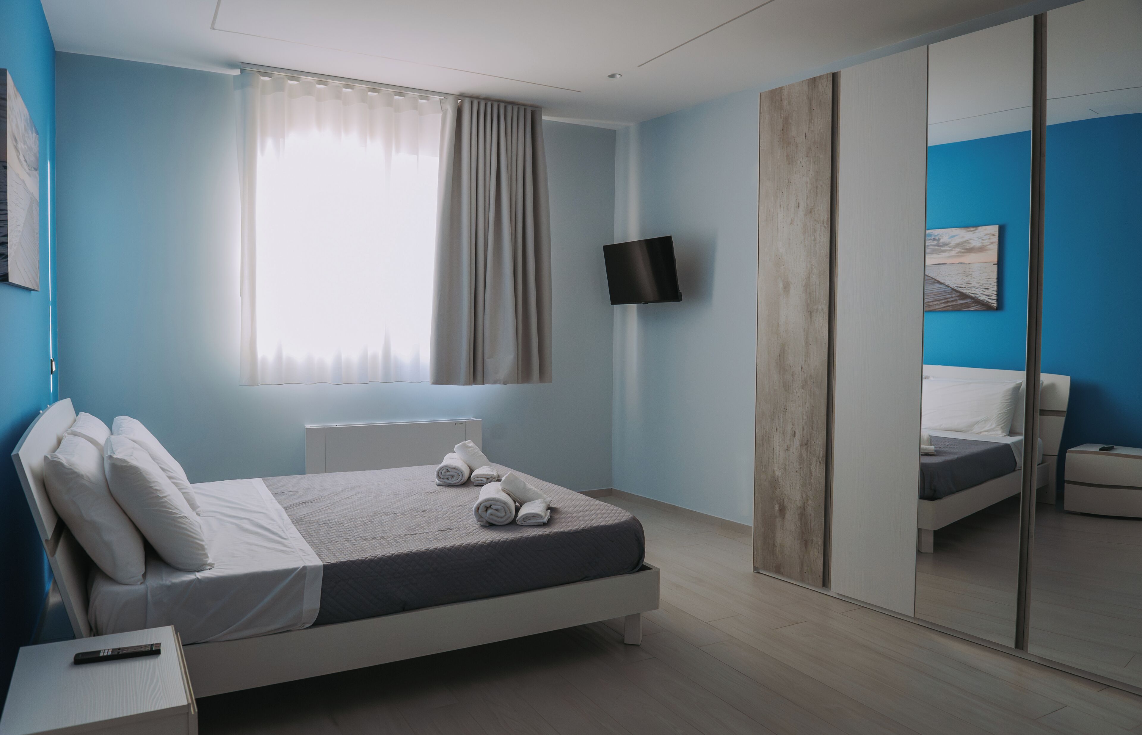 Una camera da letto moderna con un letto, una TV a parete, uno specchio grande e una finestra con tende.