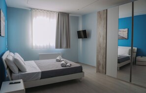 A modern bedroom with a bed, a TV mounted on the wall, a large mirror, and a window with curtains.