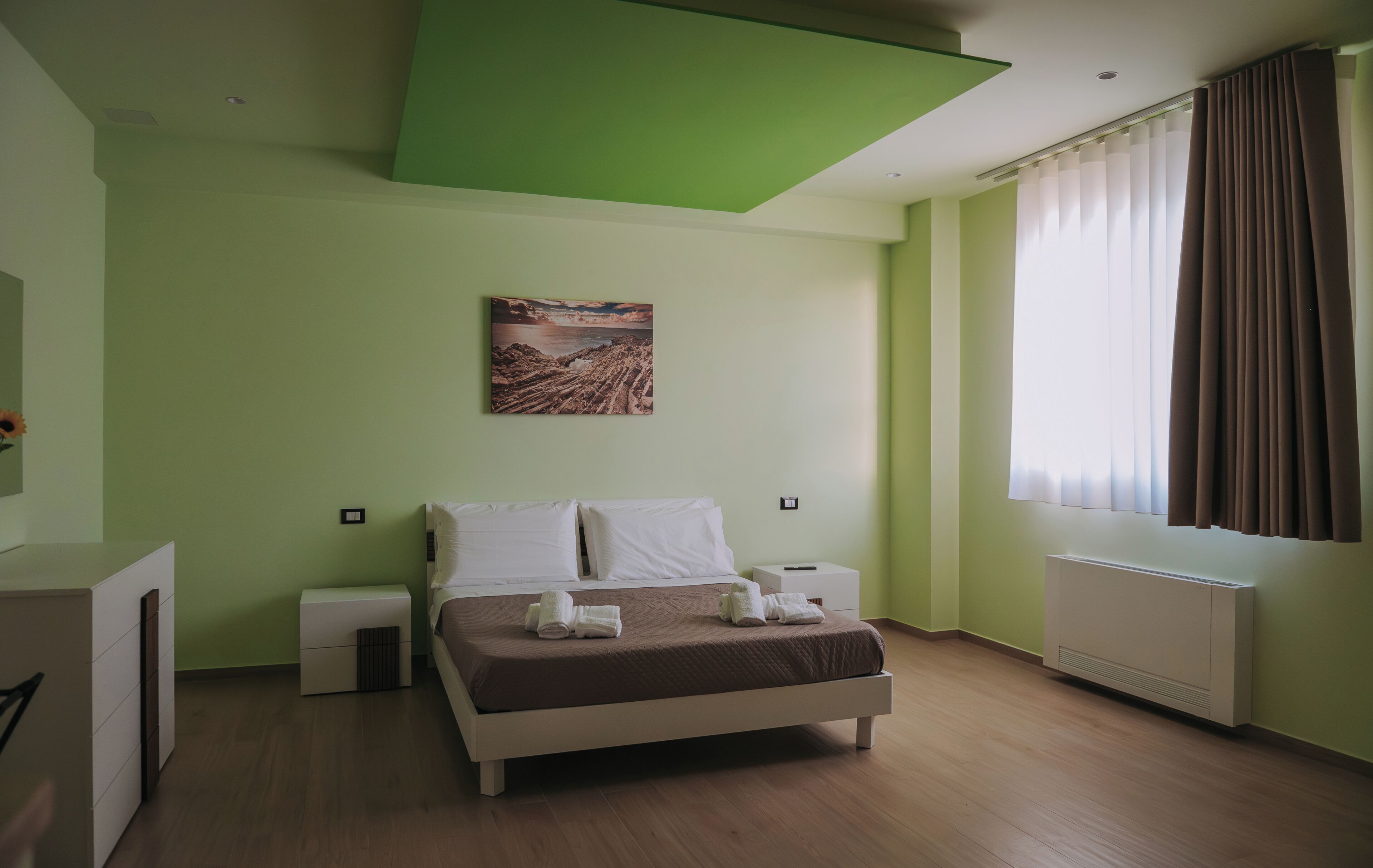 A bedroom with a bed, bedside tables, a radiator, a window with curtains, and a painting on the wall.