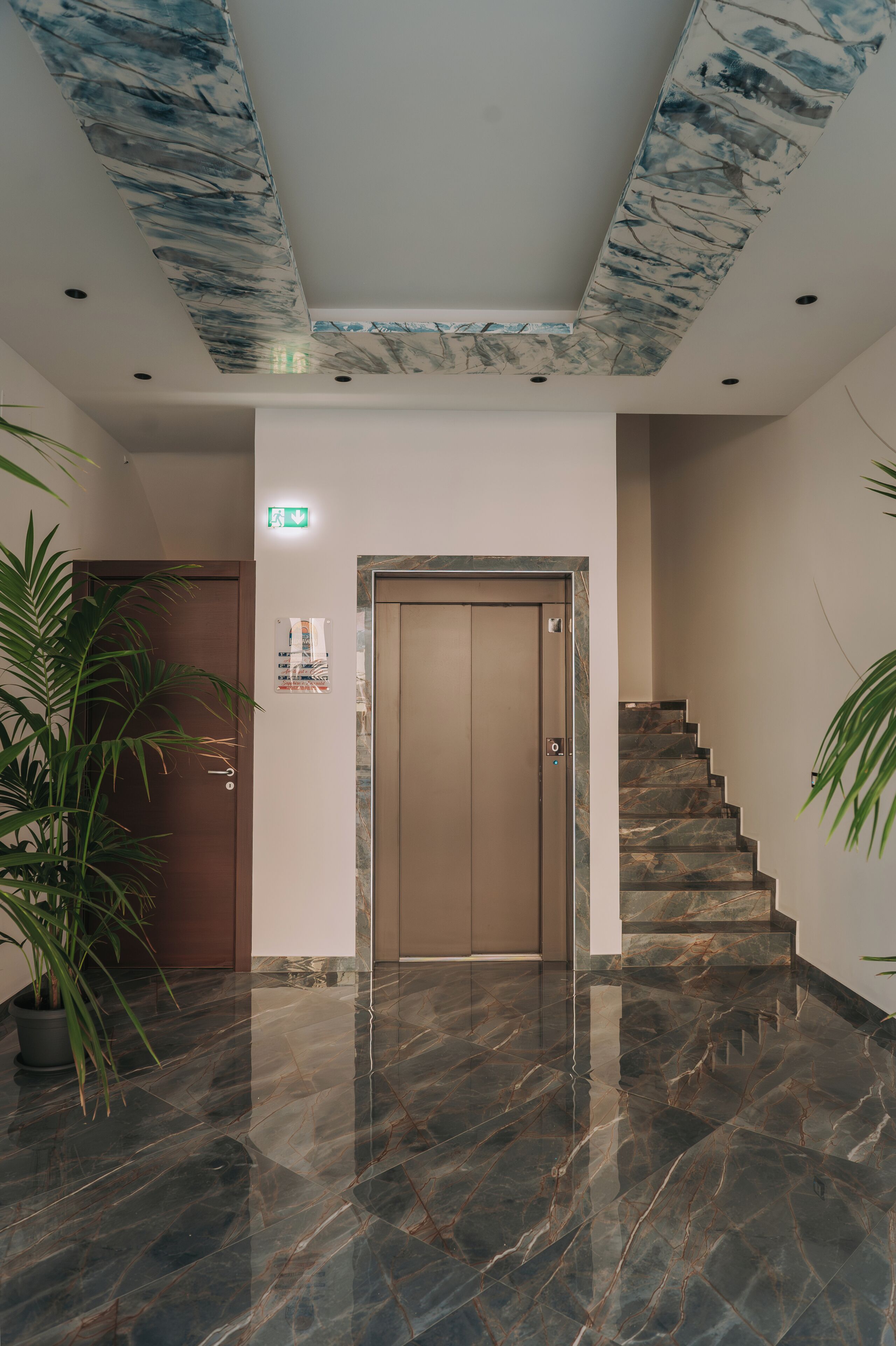 Una hall con pavimento in marmo, una porta in legno, una scalinata e un soffitto decorato con un motivo blu e bianco.