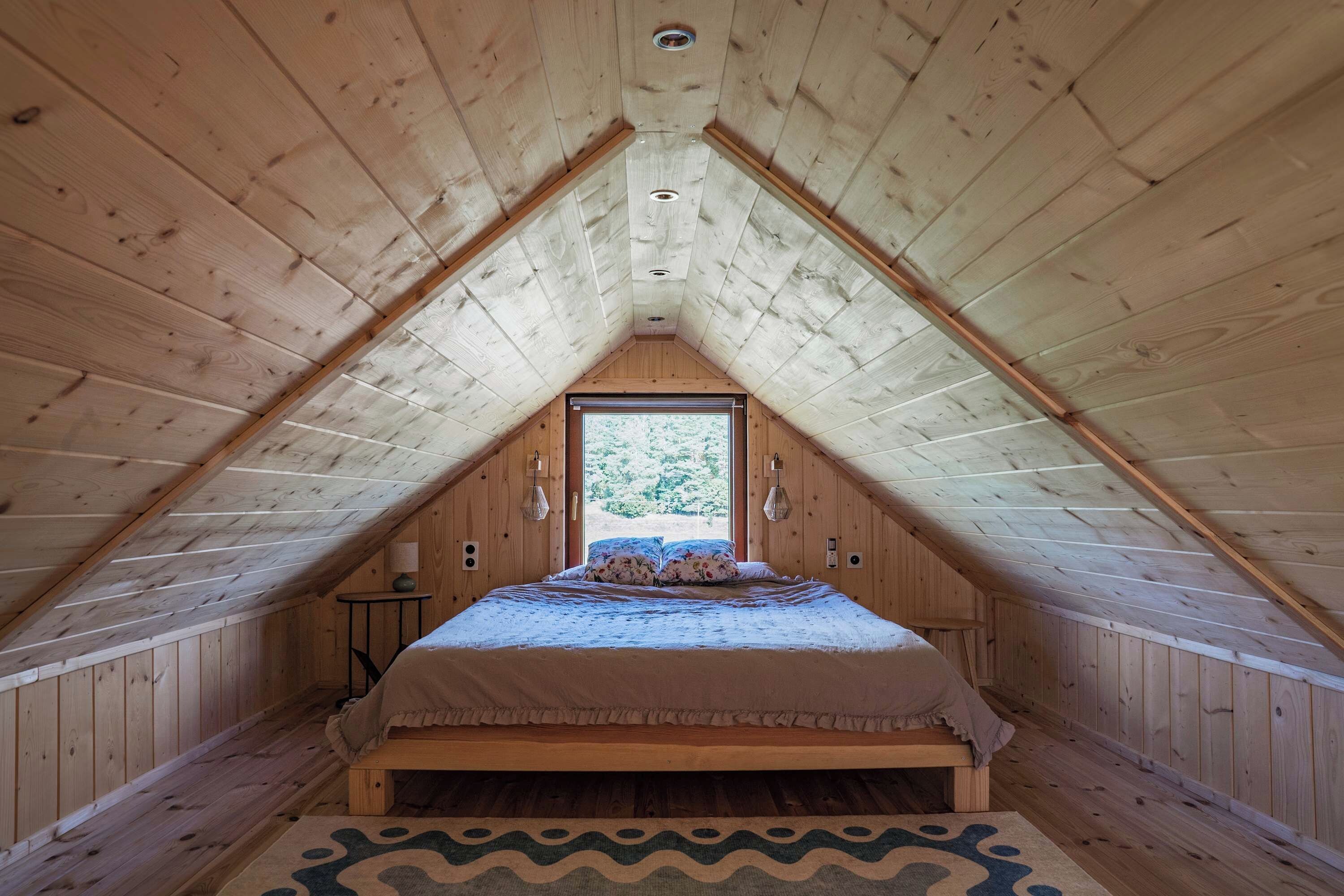 A high-ceilinged attic bedroom with a sloped roof, a bed with a floral bedspread, a small table, and a window with a view of greenery.