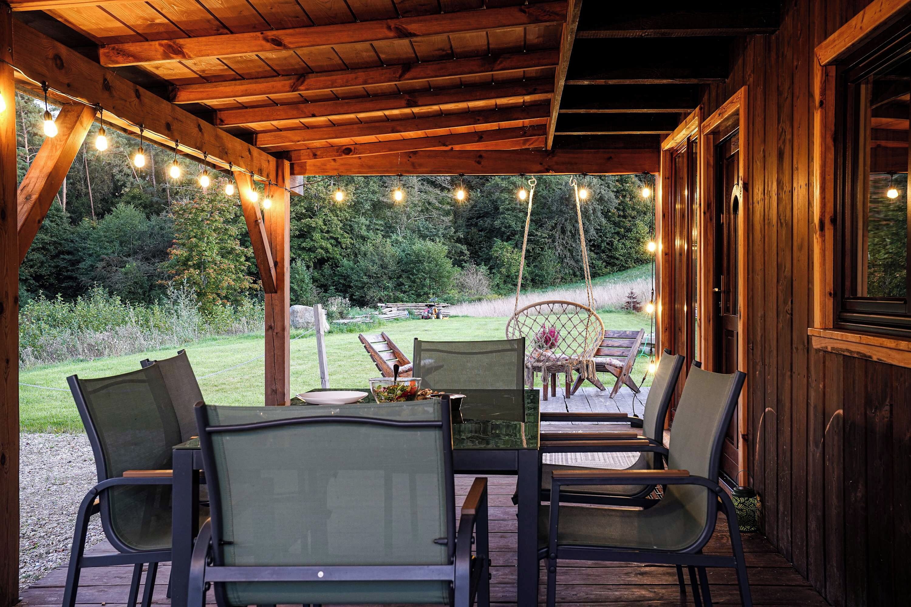 A covered outdoor seating area with wooden tables and chairs, string lights, and a view of a grassy landscape.