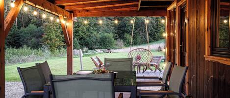 A covered outdoor seating area with wooden tables and chairs, string lights, and a view of a grassy landscape.