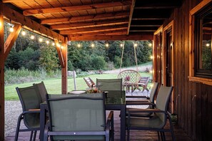 A covered outdoor seating area with wooden tables and chairs, string lights, and a view of a grassy landscape.
