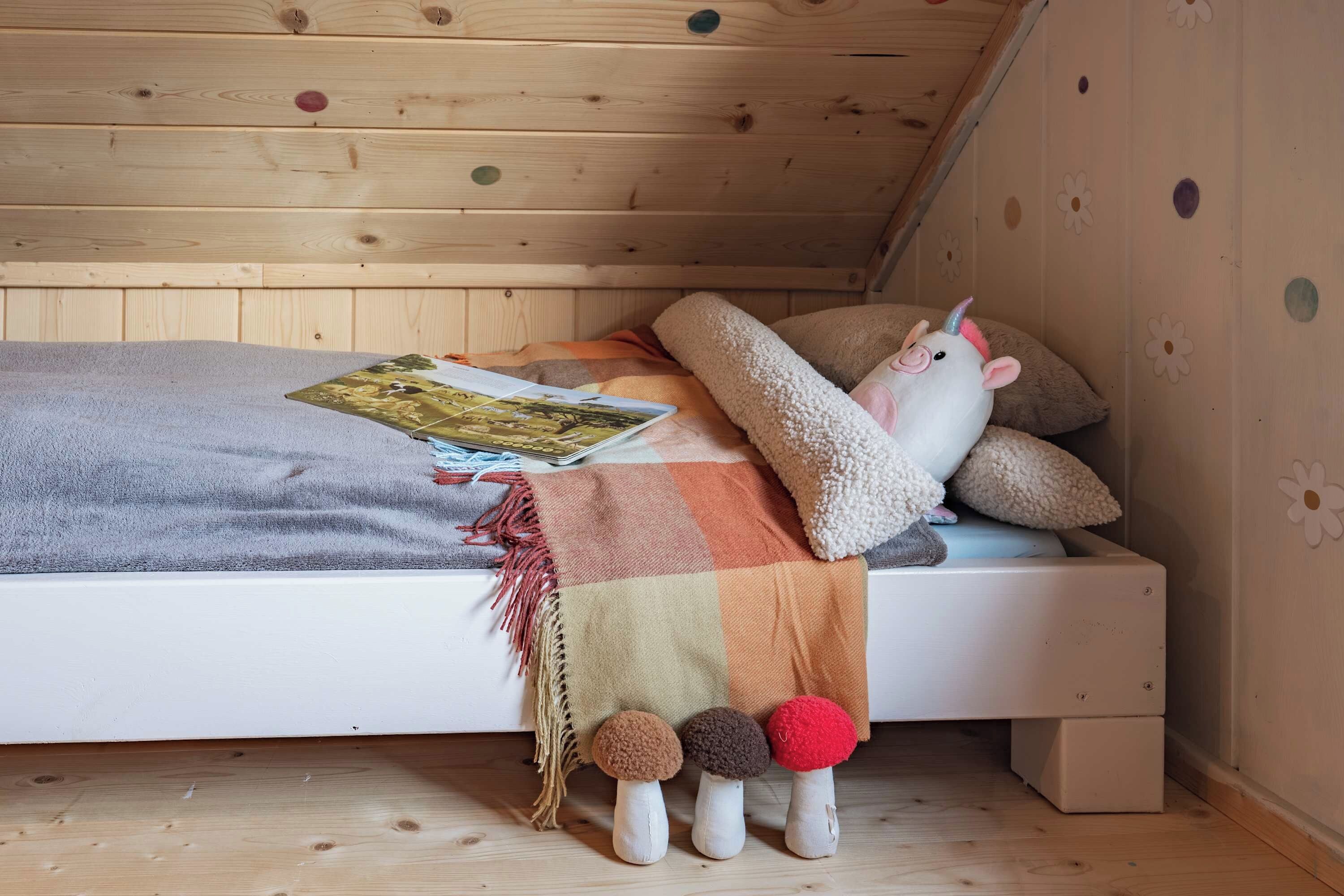 A cozy bedroom with a wooden ceiling, a bed with a blanket, pillows, a stuffed unicorn toy, and a book.