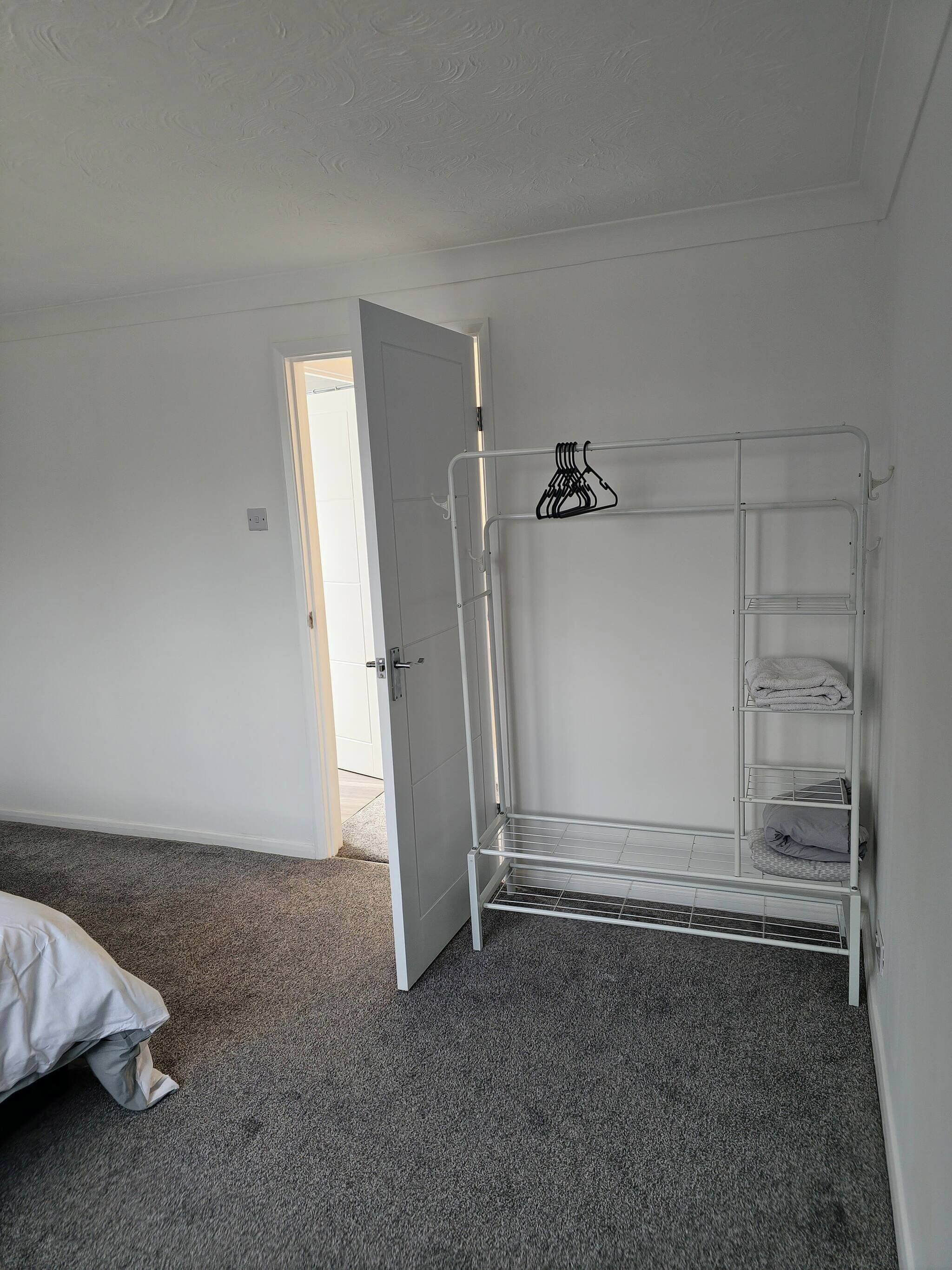 Una stanza con un mobiletto a mensole in metallo bianco appoggiato al muro, una porta che dà su un'altra stanza e un pavimento rivestito di moquette grigia.