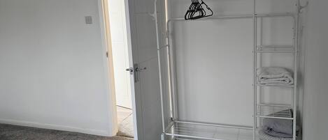 Una stanza con un mobiletto a mensole in metallo bianco appoggiato al muro, una porta che dà su un'altra stanza e un pavimento rivestito di moquette grigia.