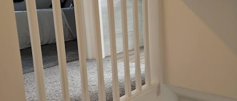 Una escalera con barandilla blanca, piso de madera, bote de basura negro y puerta de madera oscura.