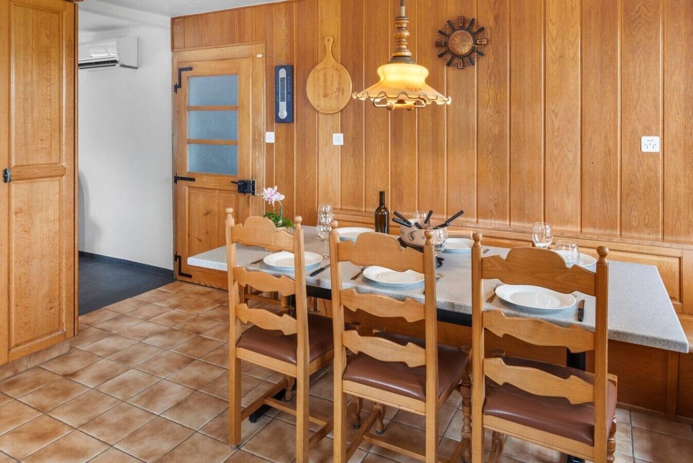 Uma sala de jantar com painéis de madeira, mesa posta para quatro pessoas, cadeiras, luminária pendente e relógio na parede.