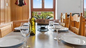 Una mesa de comedor con platos blancos, copas de vino y una botella de aceite de oliva. Hay una silla de madera con respaldo curvo y una ventana con vistas a follaje.