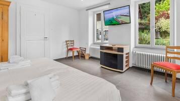 A hotel room with a bed, a TV, a desk, and a window with a view of greenery.