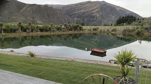 A tranquil lake with a boat, surrounded by a well-maintained lawn and a mountain backdrop. - Spa Pool | Private Pocket lake| Mountain Views,3 mins to Cromwell,walking track
(Cromwell)