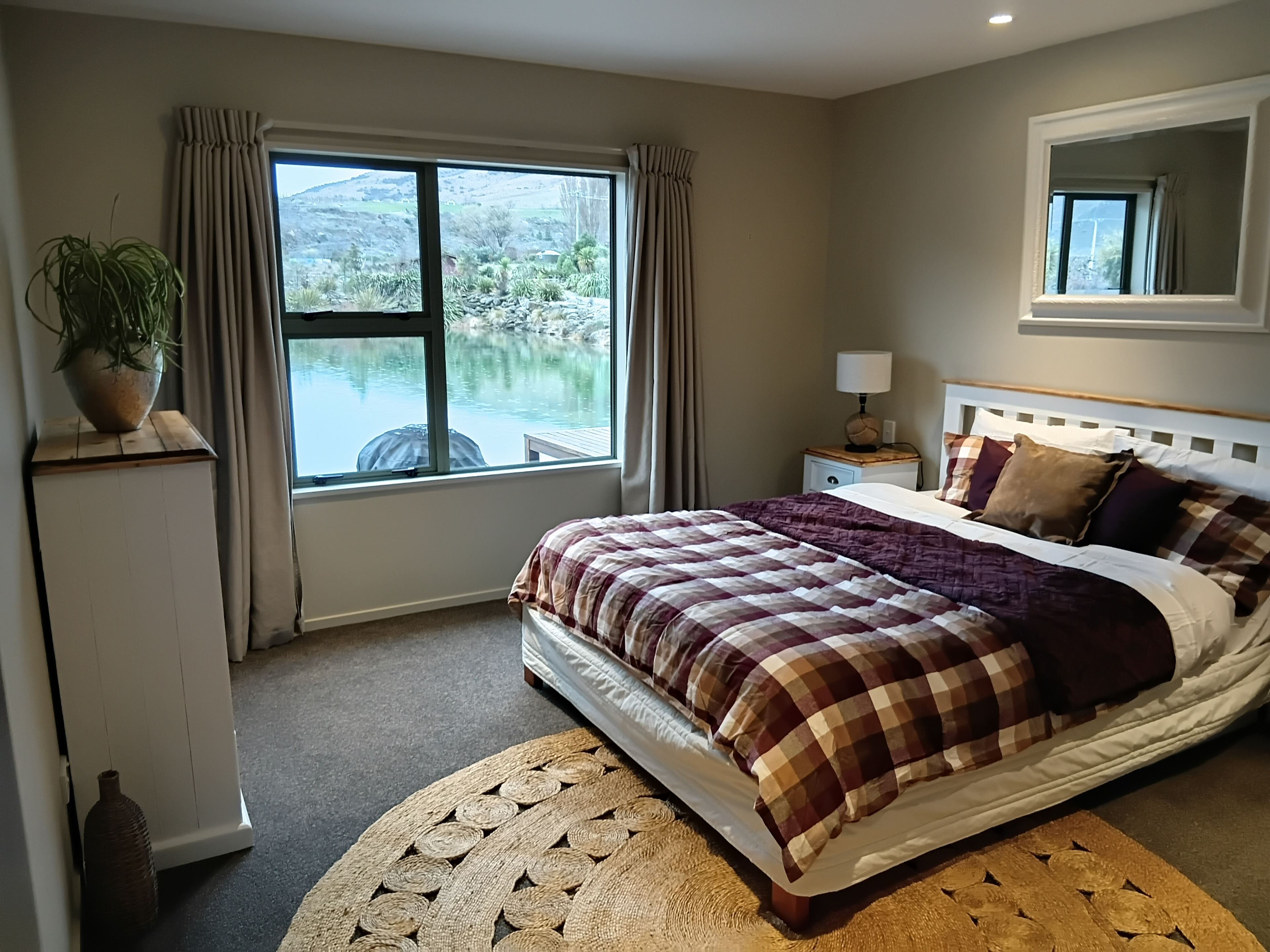 Quarto com cama, mesa de cabeceira, luminária, planta em vaso e janela com vista para um lago.