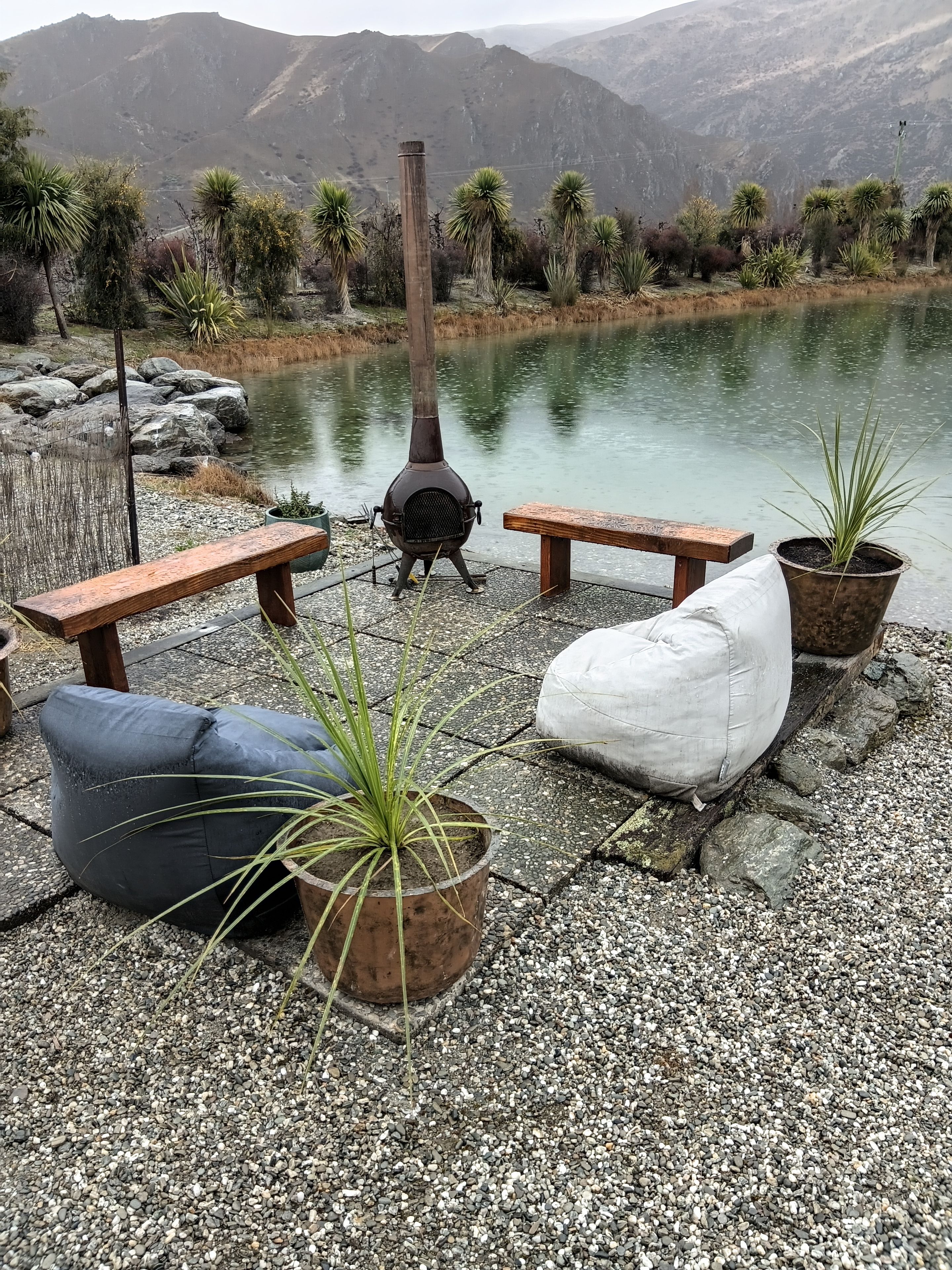 A lakeside setting with a wood-burning stove, benches, and potted plants.