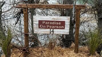 A wooden sign with the text 'Paradise on Pearson 14' in a rural setting with a body of water and trees in the background.