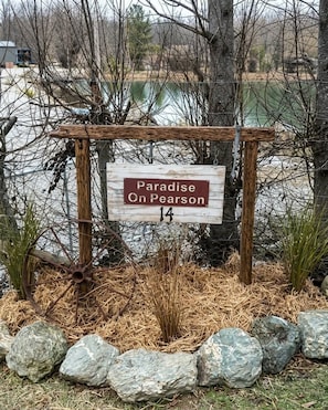 Un letrero de madera con el texto 'Paradise on Pearson 14' en un entorno rural, con un cuerpo de agua y árboles al fondo.