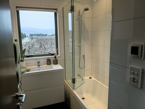 A bathroom with a bathtub, a shower, a window with a view of a cityscape, and a sink with two bottles. - Paradise  on Pearson,Spa Pool | Private Pond | Mountain Views,3 mins to Cromwell (Cromwell)