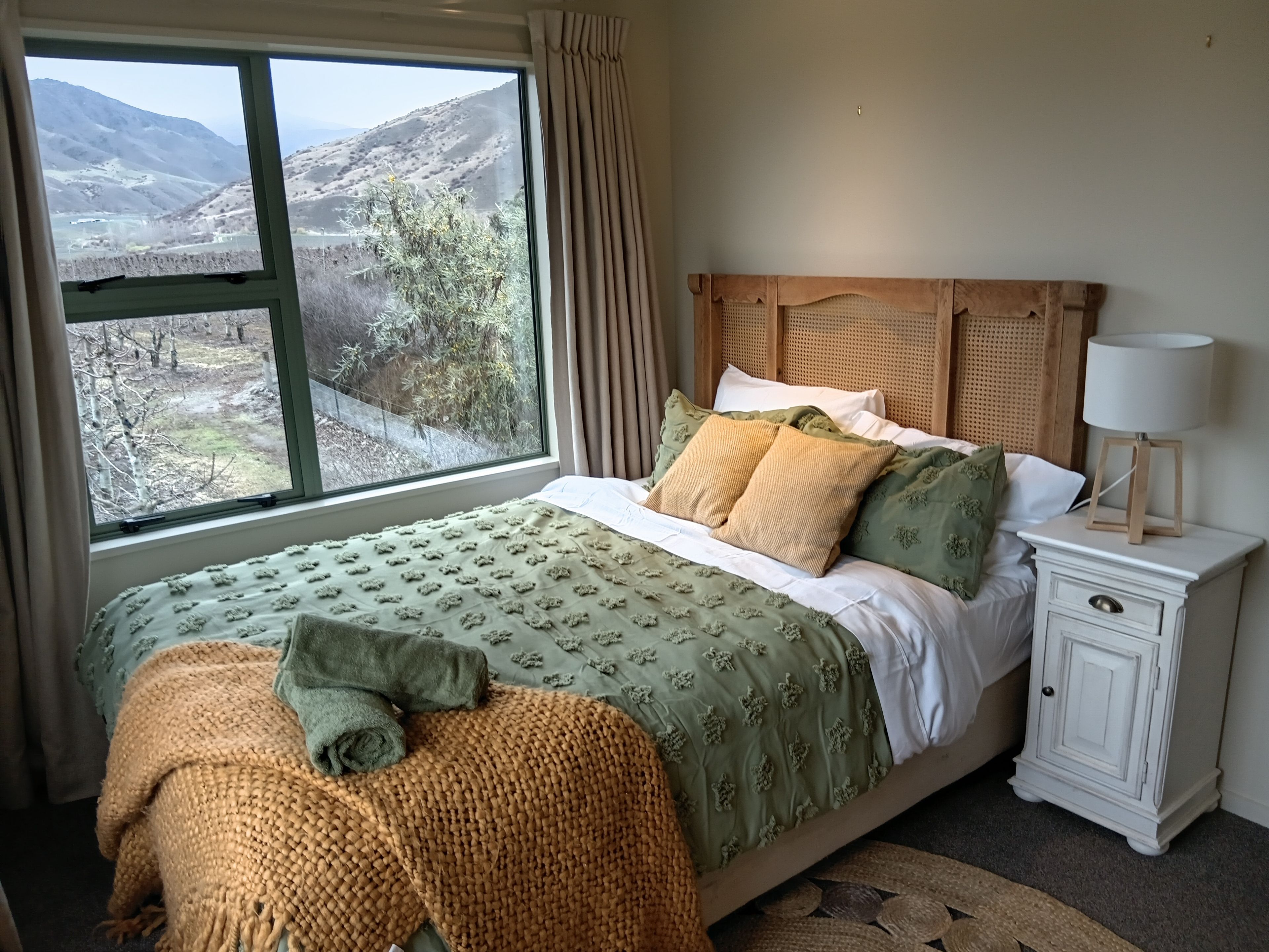 A bedroom with a large window, a bed with green and white bedding, a wooden headboard, a bedside table with a lamp, and a view of a mountainous landscape.