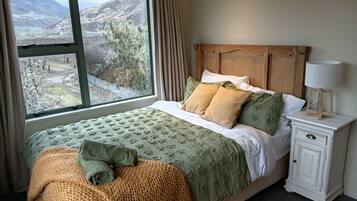 A bedroom with a large window, a bed with green and white bedding, a wooden headboard, a bedside table with a lamp, and a view of a mountainous landscape.