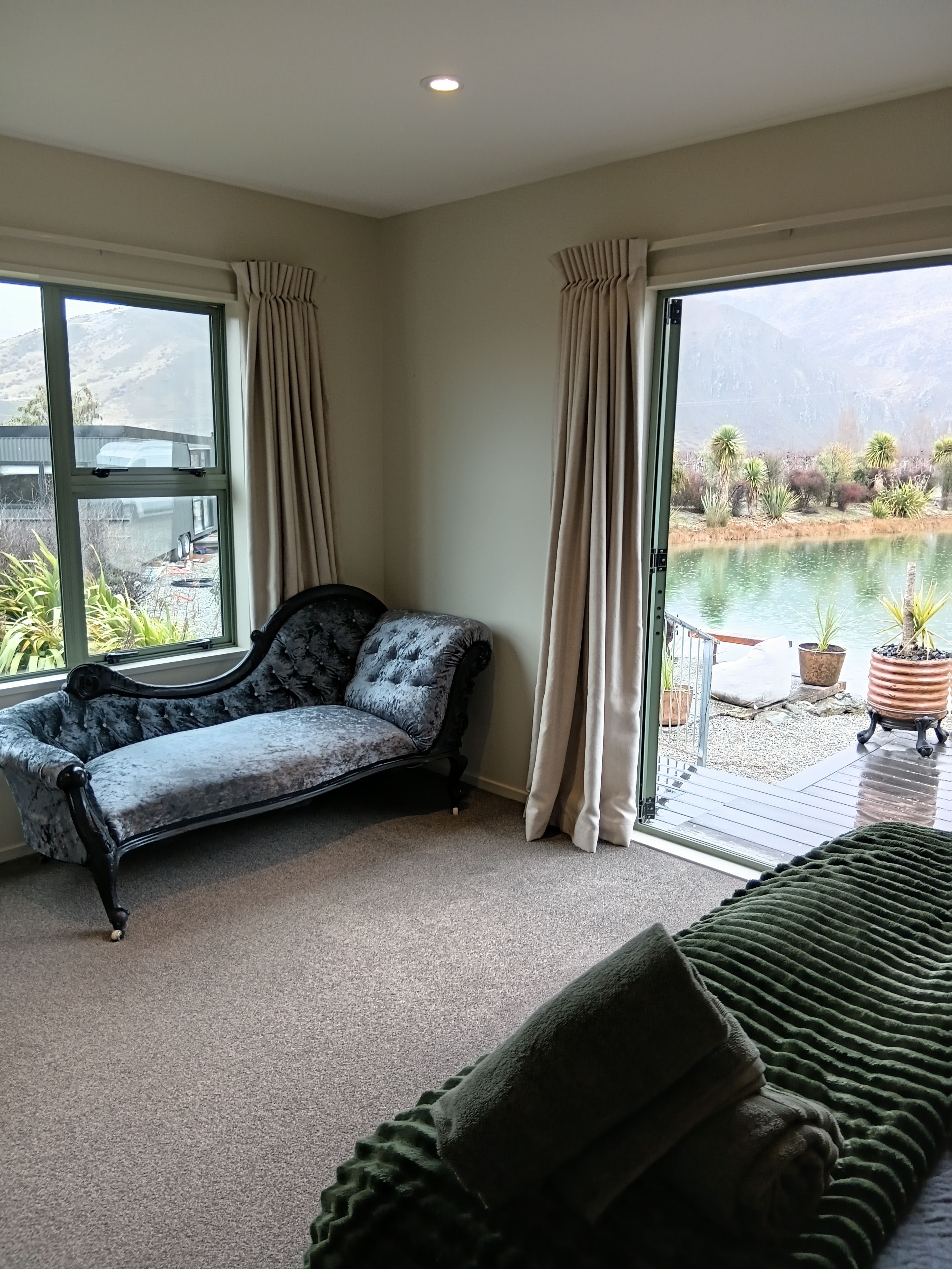 Quarto com chaise longue, manta verde e vista para um lago através de amplas janelas.