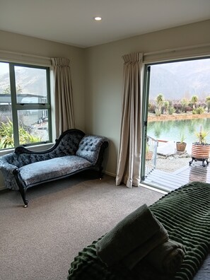 Habitación con una chaise longue, una manta verde y vista a un lago a través de amplios ventanales.