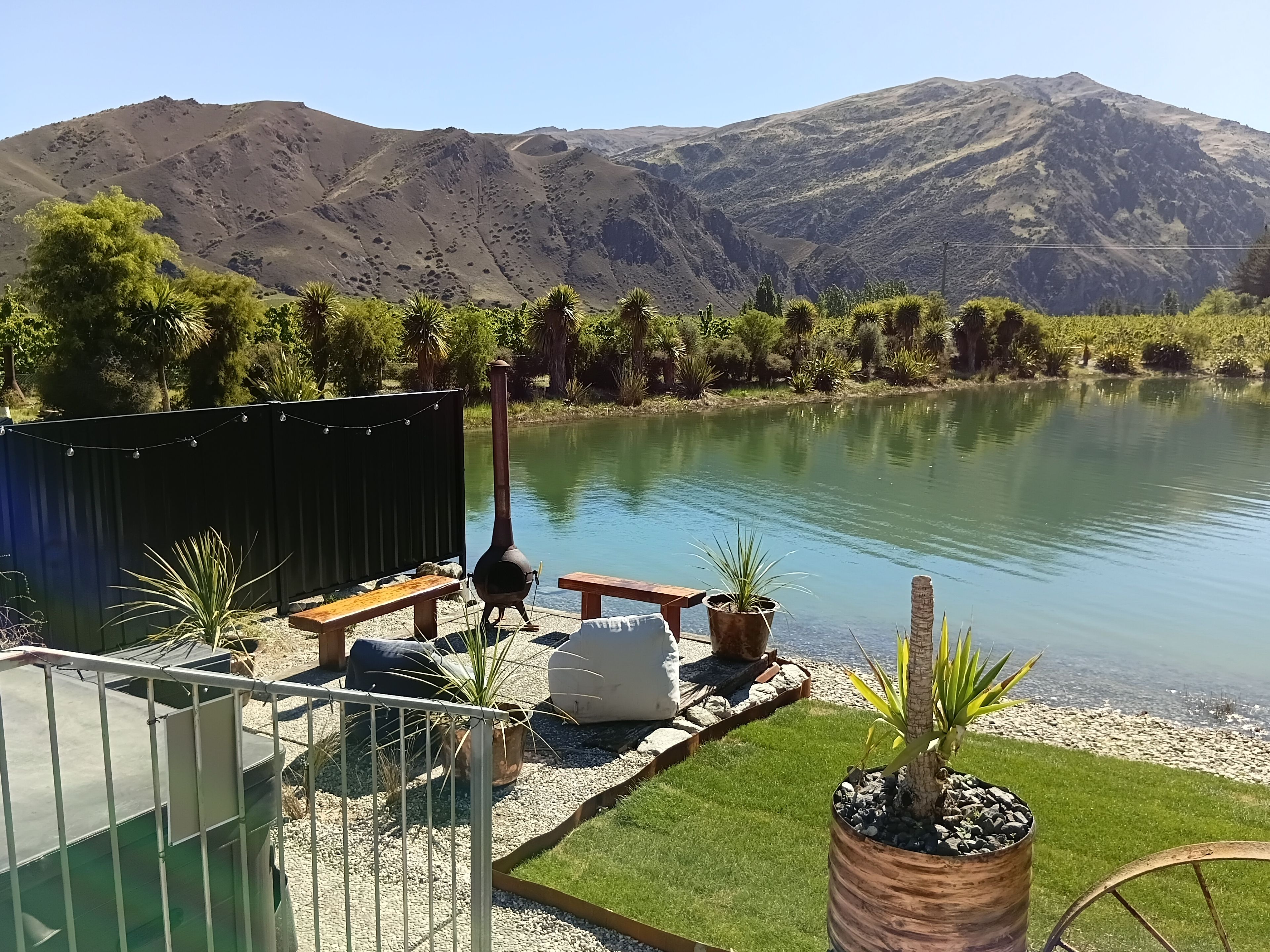 A lakeside patio with a wooden bench, a metal grill, and potted plants.