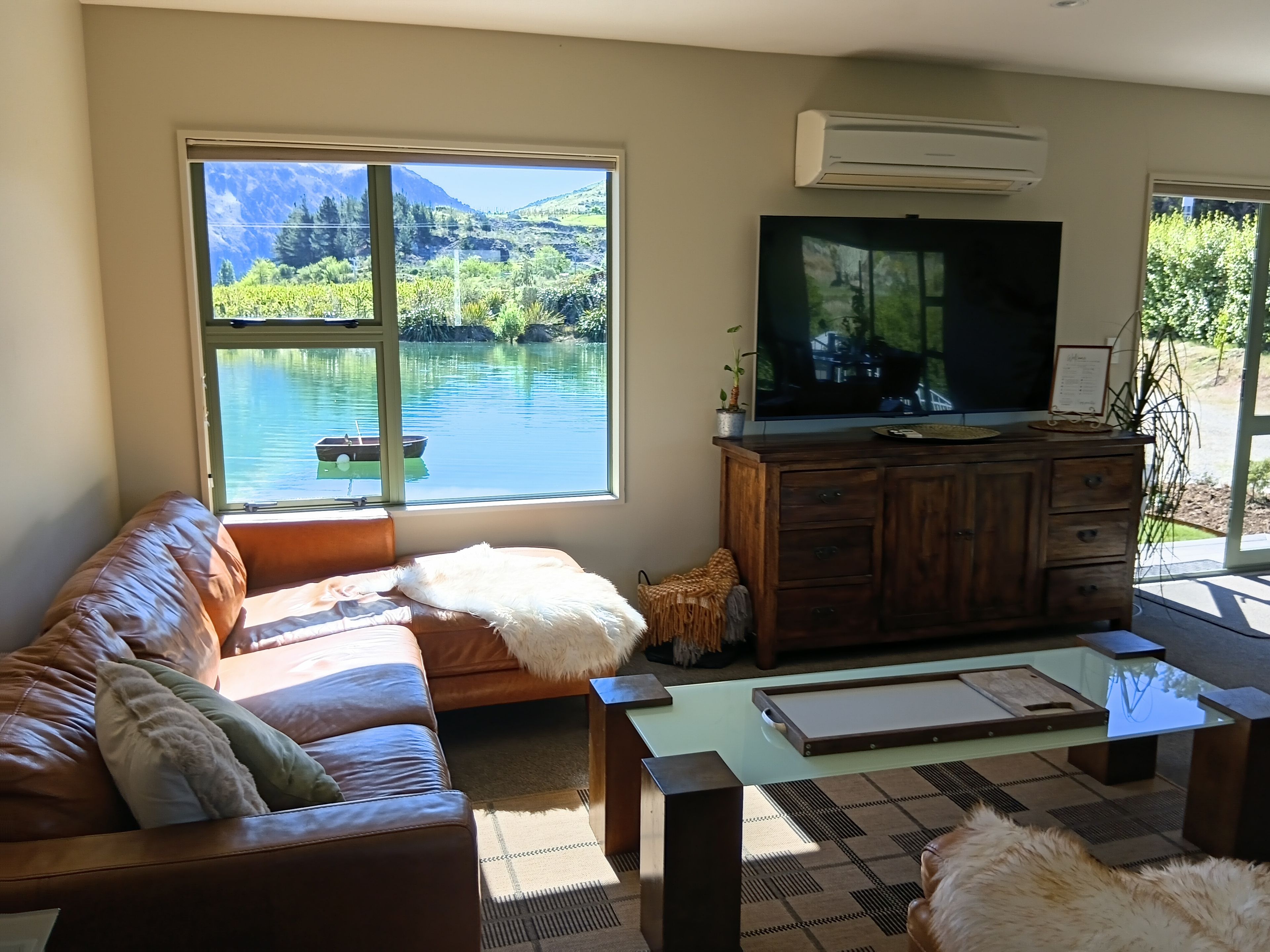 A living room with a large window overlooking a lake, a television, and a wooden cabinet.