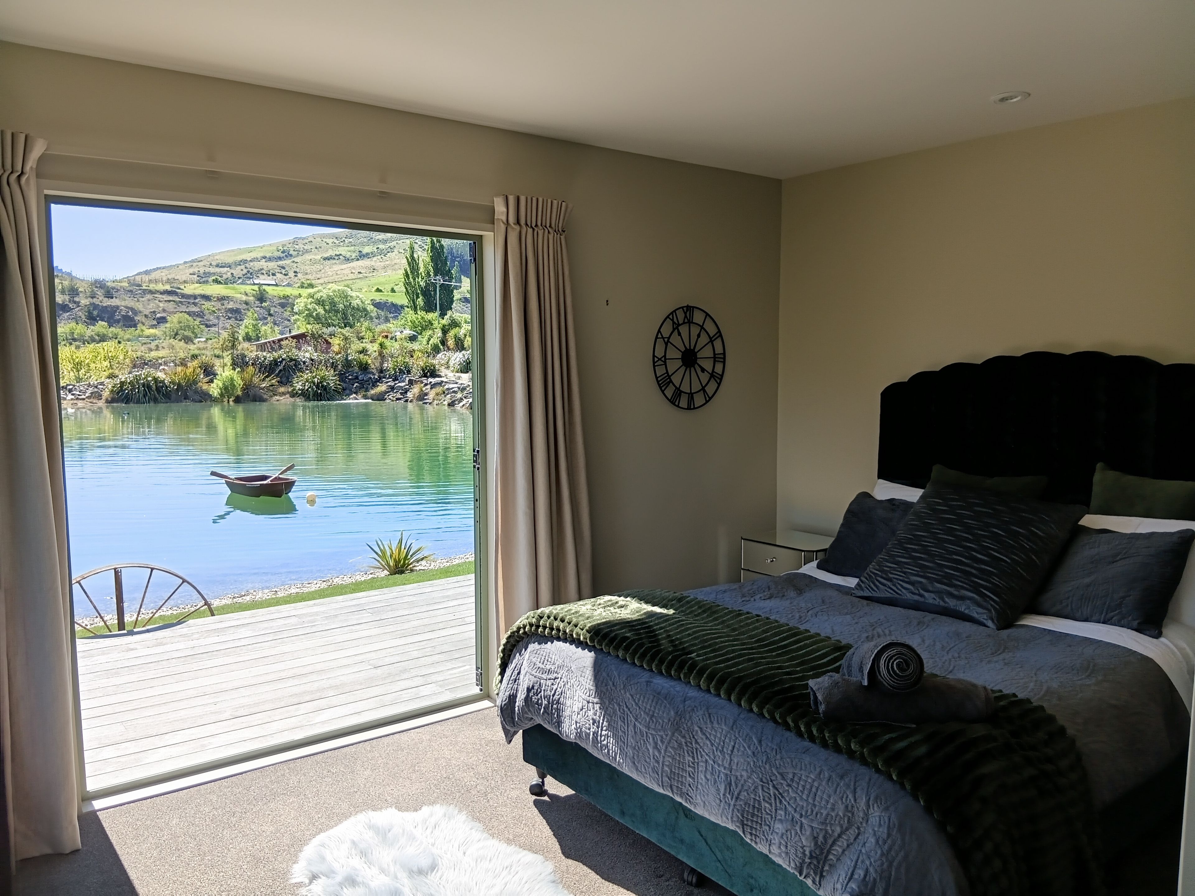 A bedroom with a large window overlooking a lake, a bed with a dark headboard, and a wall clock.