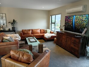 A living room with a brown leather sofa, a coffee table, a TV, and a large window. - Paradise  on Pearson,Spa Pool | Private Pond | Mountain Views,3 mins to Cromwell (Cromwell)