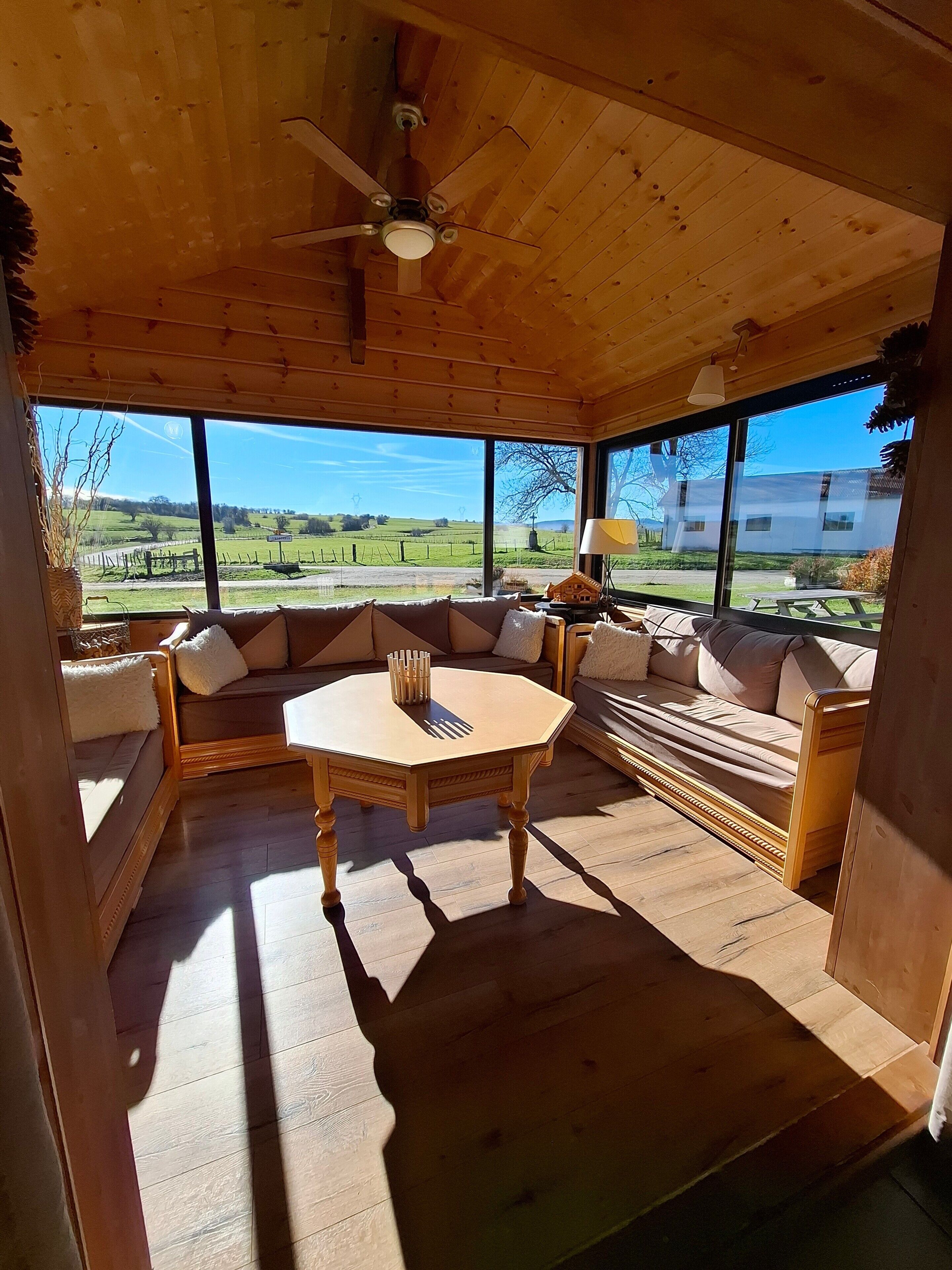 A wooden interior with a round table, sofas, and large windows offering a view of the countryside.