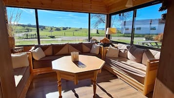 A wooden interior with a round table, sofas, and large windows offering a view of the countryside.