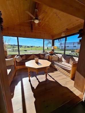Interior de madera con una mesa redonda, sofás y ventanas grandes que dan a la campiña.