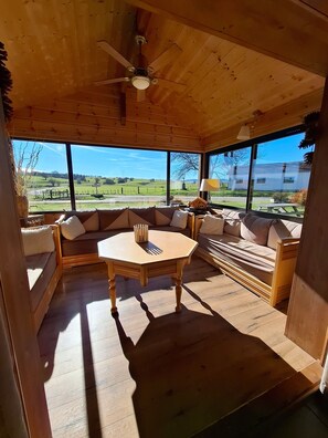 A wooden interior with a round table, sofas, and large windows offering a view of the countryside.