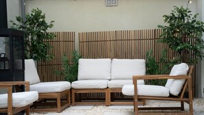 Un área de estar al aire libre con madera, cojines blancos, una valla de bambú y plantas en macetas.