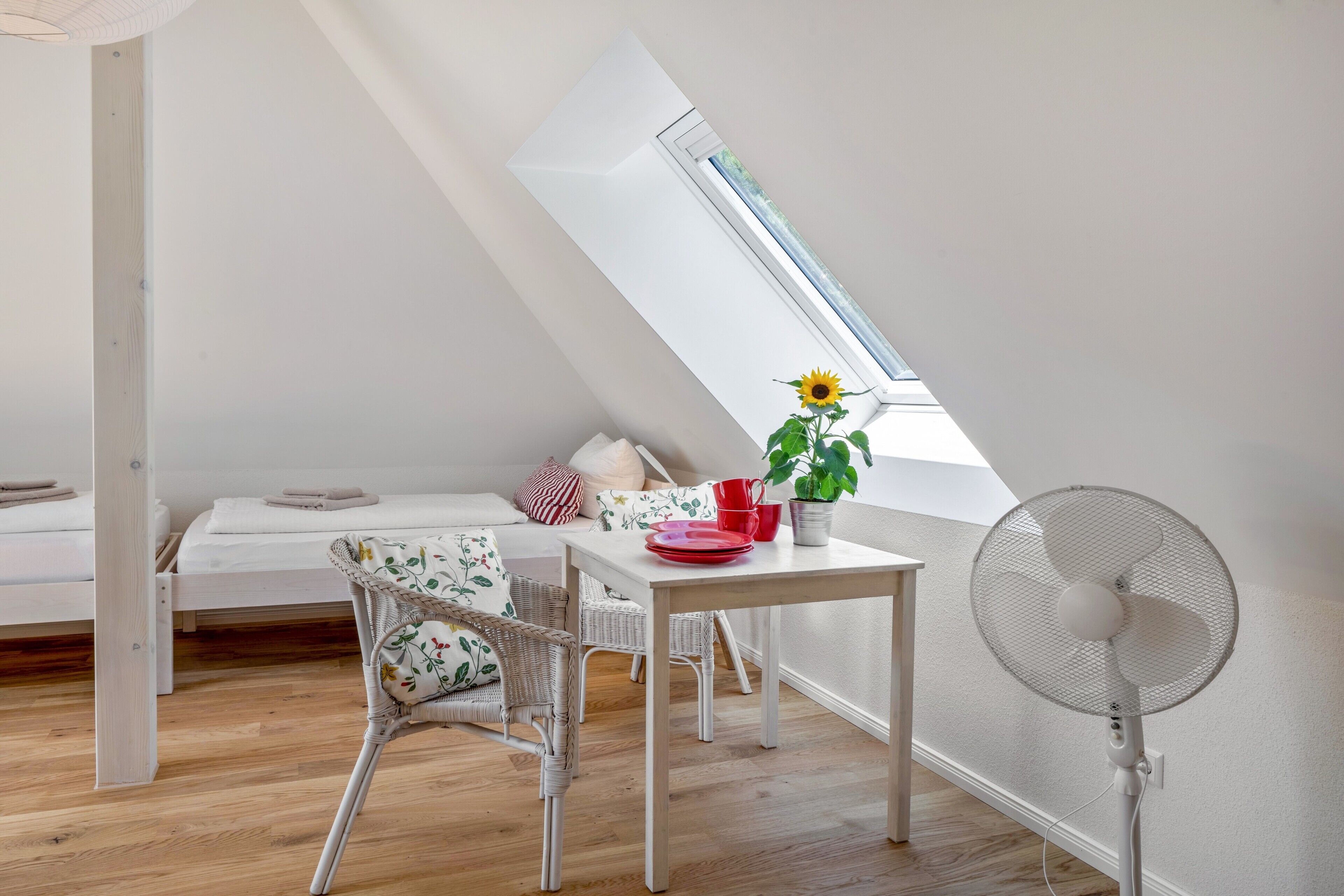 A small, cozy room with a sloped ceiling, a bed, a dining table with chairs, a potted plant, and a standing fan.