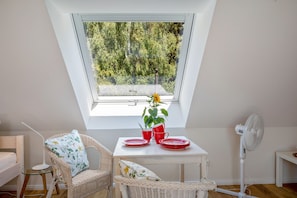 A small, bright room with a sloped ceiling, a table with red plates and a potted plant, wicker chairs, and a standing fan. - Ferienwohnung 'Fuchs und Has Feldberg-Neu Eröffnung Juli 2026 - App 8' mit toller Aussicht, Gemeinschaftsgarten und WLAN (Feldberg)