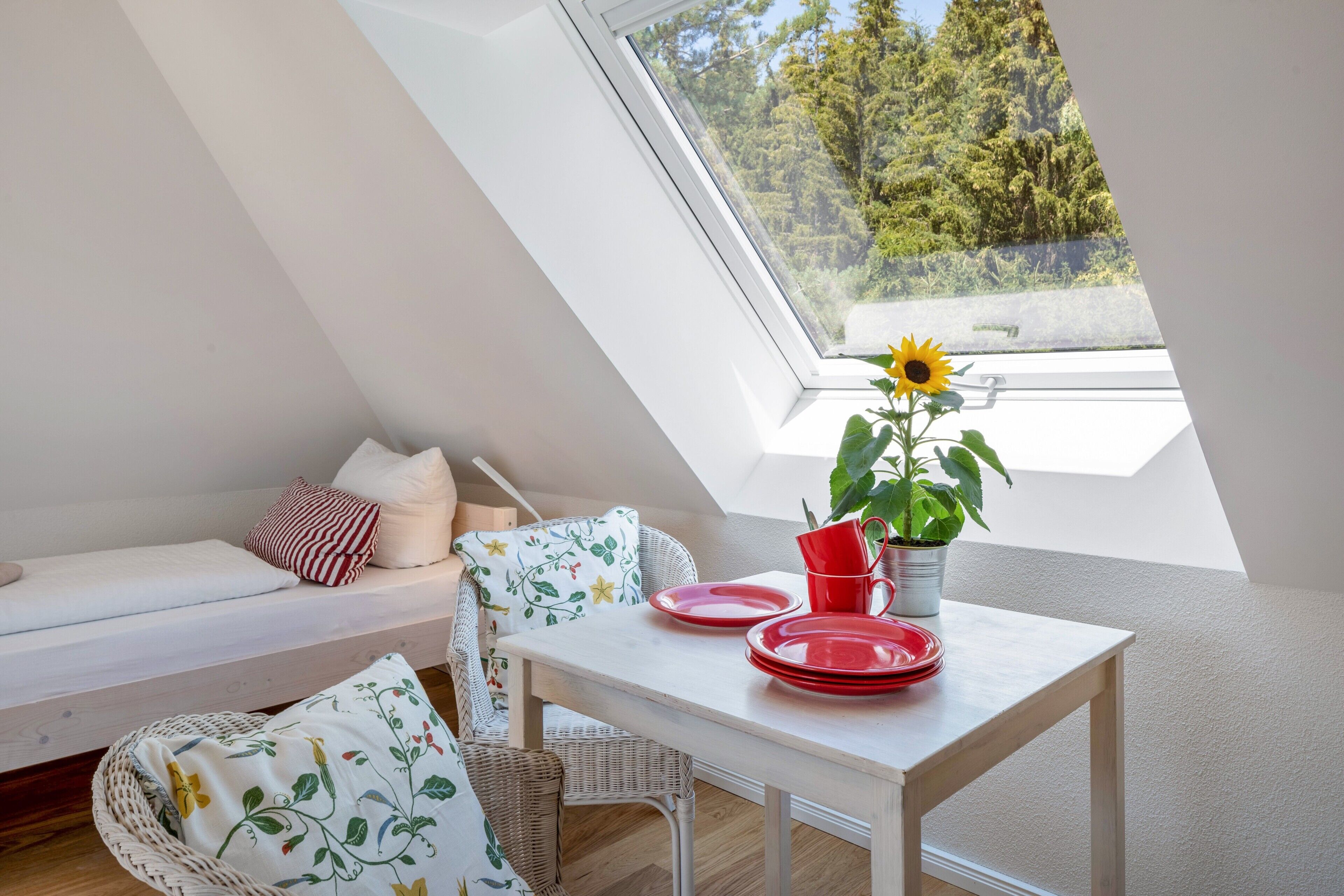 A room with a sloped ceiling, a dining table with red plates, a sunflower, and a view of trees through a skylight.