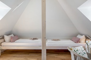 Ein Schlafzimmer im Loft-Stil mit schrägem Dach, Holzbalken, einem Bett mit weißer Bettwäsche und gestreifteren Kissen, einem kleinen Tisch und einem Korbstuhl.