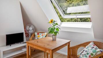 Ein Zimmer mit schräg gedachter Decke, einem Holztisch, einem Korbstuhl, einem Fernseher, einem Ventilator und einem Fenster mit Blick auf Grünflächen.