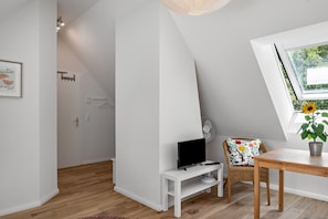 Una stanza con soffitto spiovente, una televisione su un supporto bianco, una sedia in vimini con cuscino a fiori, un tavolo di legno e una finestra con un girasole.