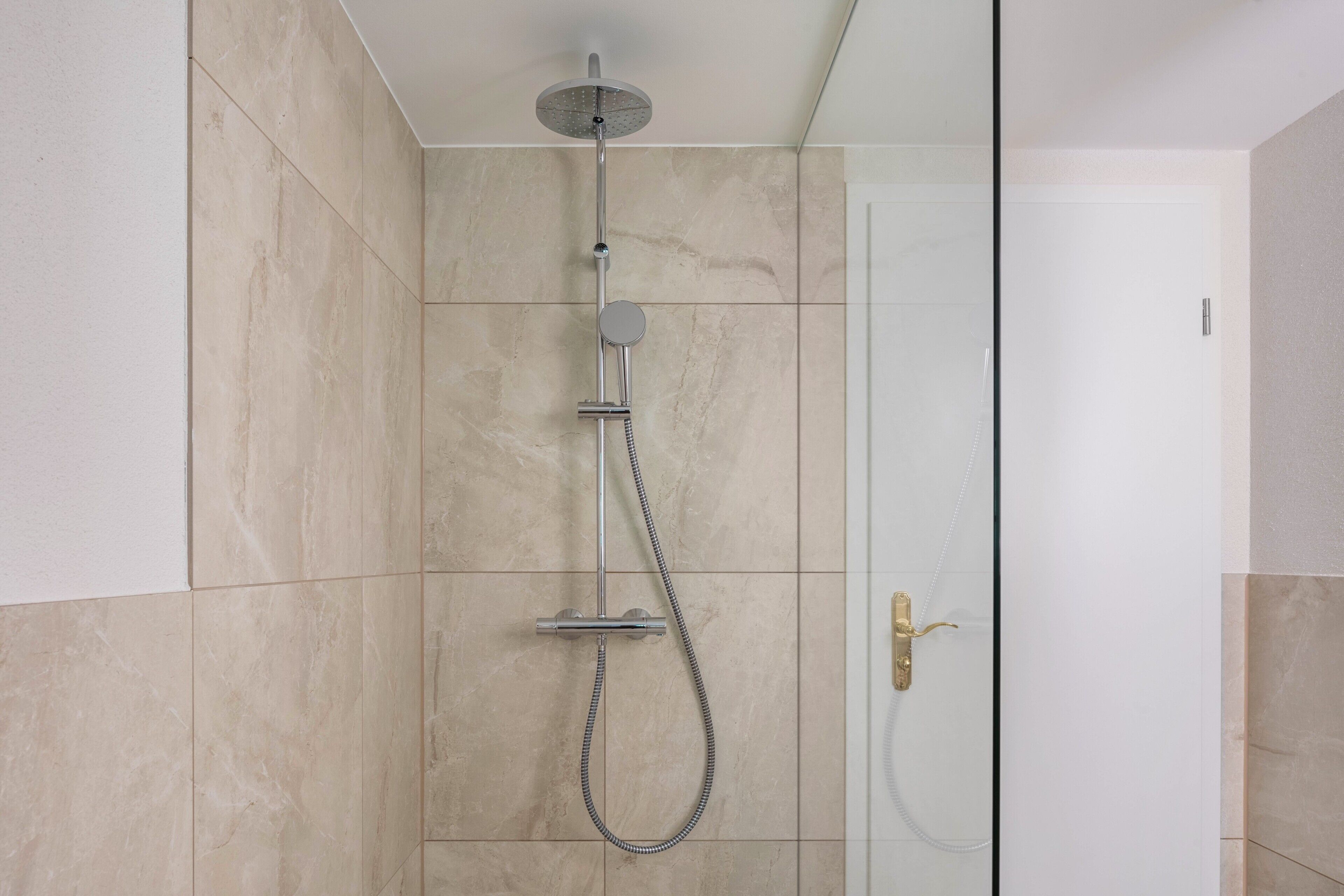 Eine Dusche mit Handbrause und Wandarmatur, eine Glastür mit goldener Griff und beige geflieste Wände.